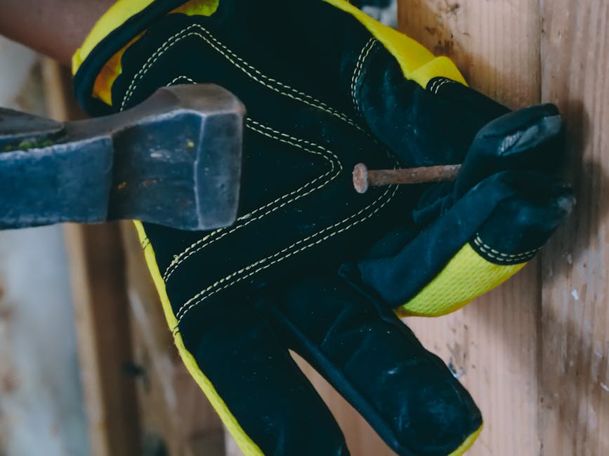 Worker using a hammer and nail on a wooden surface with protective gloves.