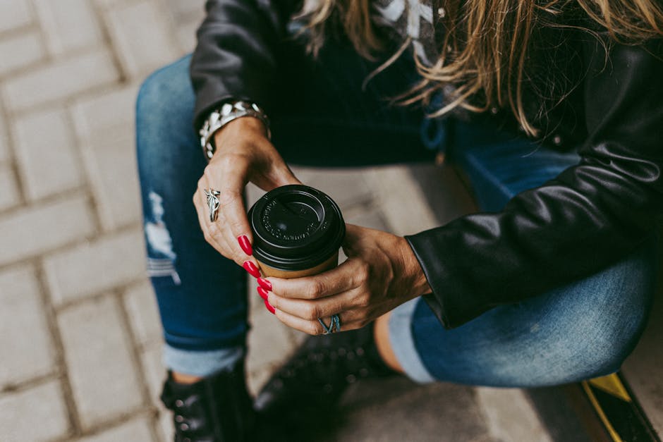 Woman in jeans and jacket enjoys coffee outdoors, leisurely sitting.