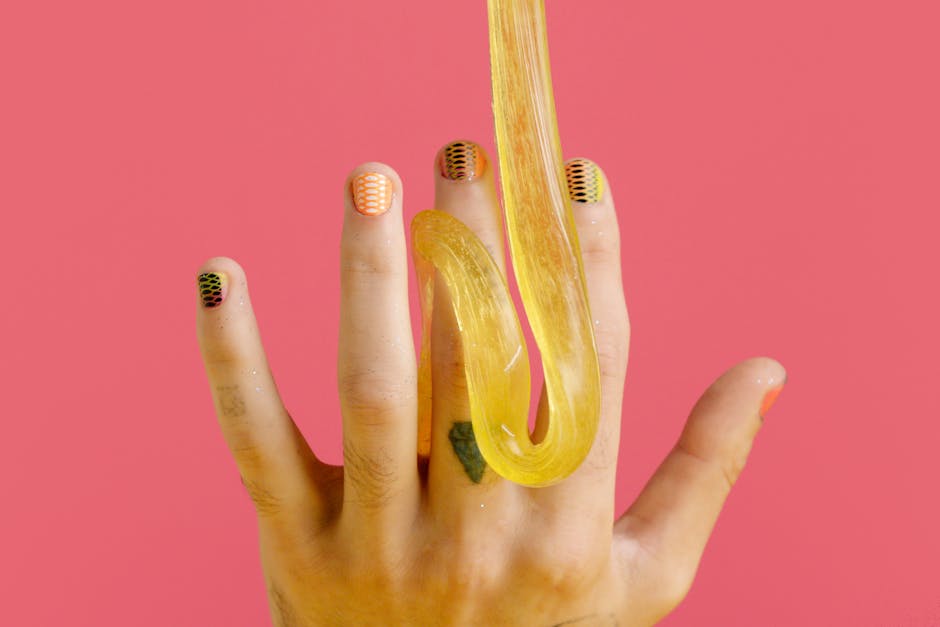 Creative nail art and yellow slime draped on fingers against a pink backdrop.