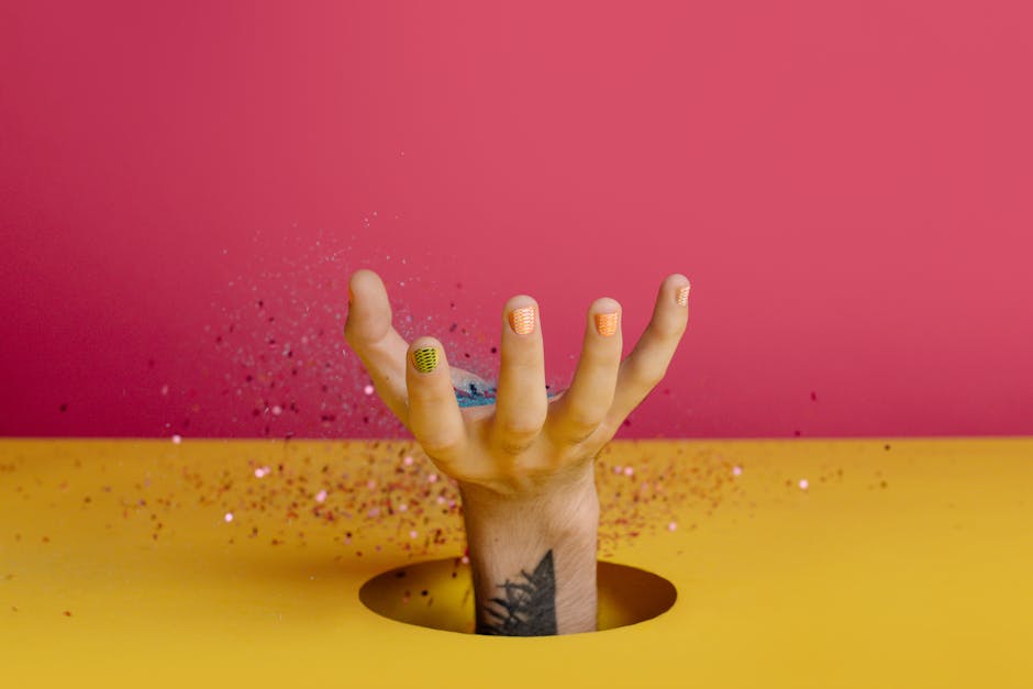 A colorful hand with manicured nails reaching up through a bright background with floating glitter.