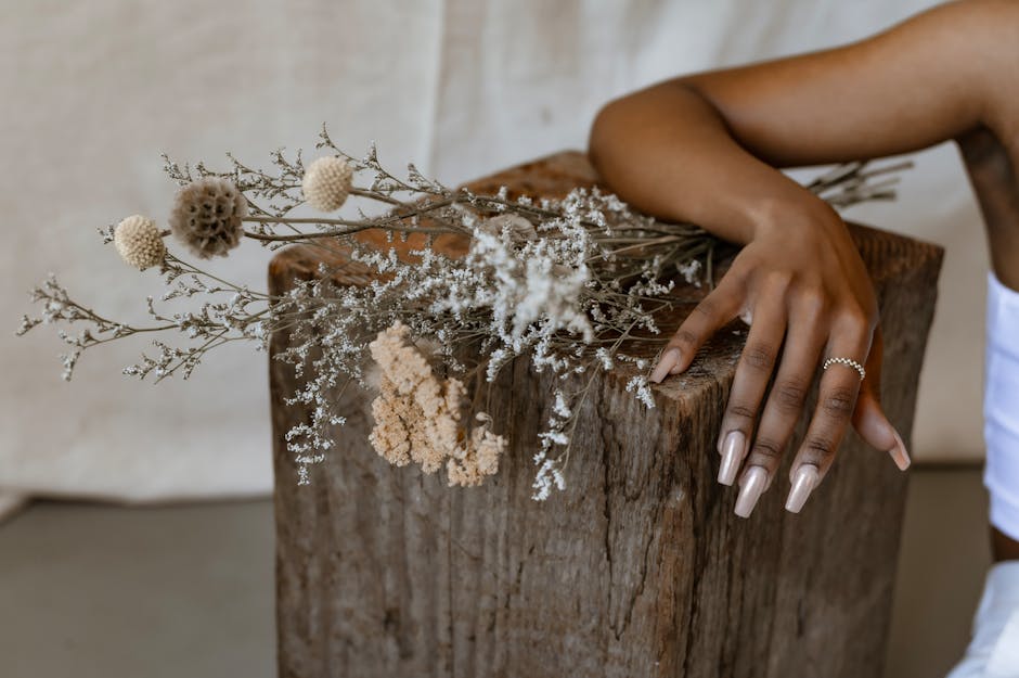A delicate hand with manicured nails and silver ring rests on a rustic wooden block adorned with dried flowers.